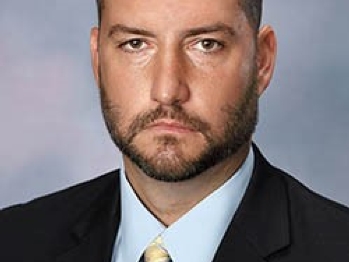 A headshot of a white man with a moustache and beard, dressed in a dark coat, white shirt, and light yellow tie/