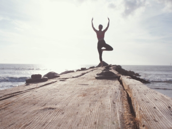 Woman standing on a pier next to the ocean in a yoga pose back lit by the sun