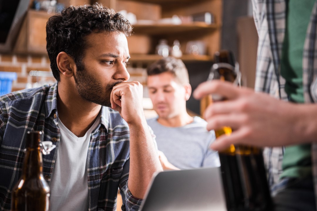Man sitting pensively with his chin in his hand at a bar while others around him drink.